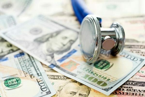 A stethoscope resting on top of scattered U.S. hundred-dollar bills. The close-up shot highlights the connection between healthcare and financial costs, symbolizing medical expenses, insurance, or healthcare funding.