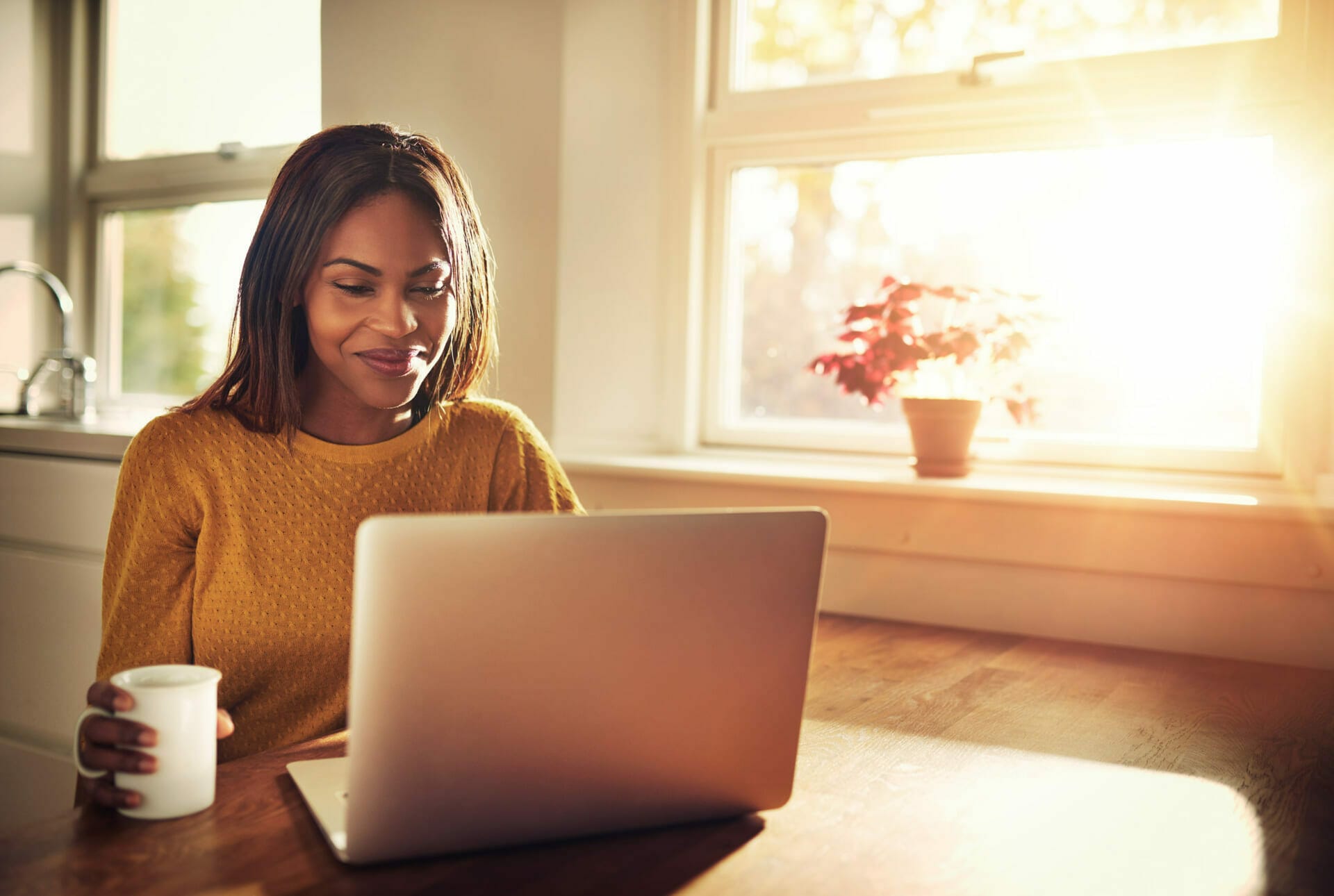easy to work with The image depicts a woman in a mustard yellow sweater, engaging with a laptop on a table while holding a mug in her hand. The indoor setting is warmly lit by sunlight streaming through a window, which also illuminates a potted red plant on the windowsill. The woman appears focused and content, suggesting a comfortable and serene work-from-home environment.