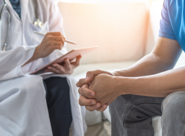 A patient sitting on the left is engaged in a discussion with a medical professional, who is holding a tablet and seems to be providing information or taking notes. The patient, clasping their hands, appears attentive and possibly anxious. This scene suggests a consultation or discussion about treatment options, possibly relating to alternative cancer treatment centers, as the context provided is a webpage about alternative cancer treatment guides.