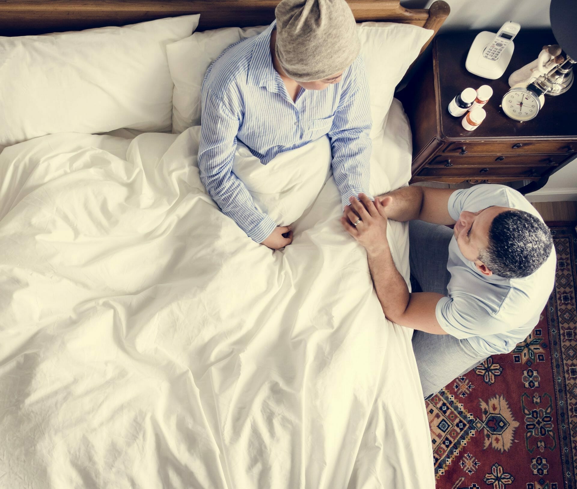 A person in a hospital bed, wearing a beanie, is being comforted by someone sitting beside them, holding their hands. There are medications on a nightstand.