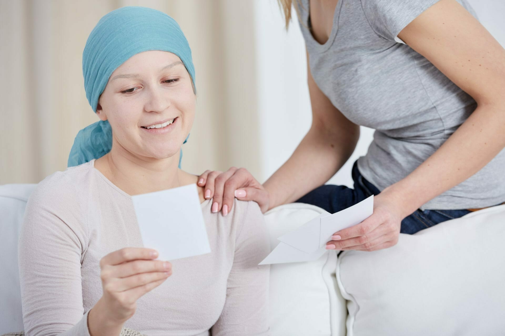 A woman wearing a headscarf, indicating cancer treatment, smiles while looking at a photo. Another woman beside her offers support with a hand on her shoulder.