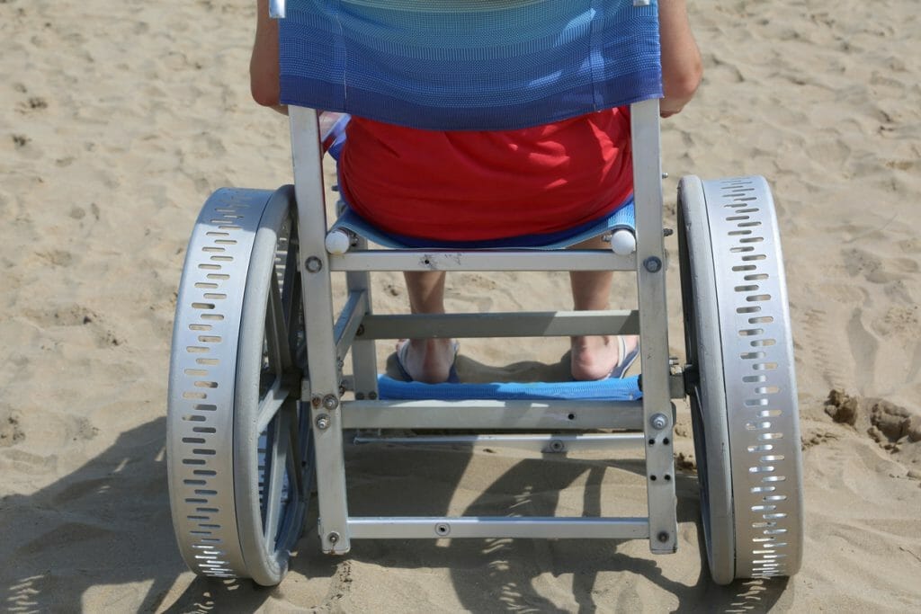 A person in a wheelchair with large, specialized wheels is on the beach, highlighting the mobility aids and associated costs for ALS treatment and care.