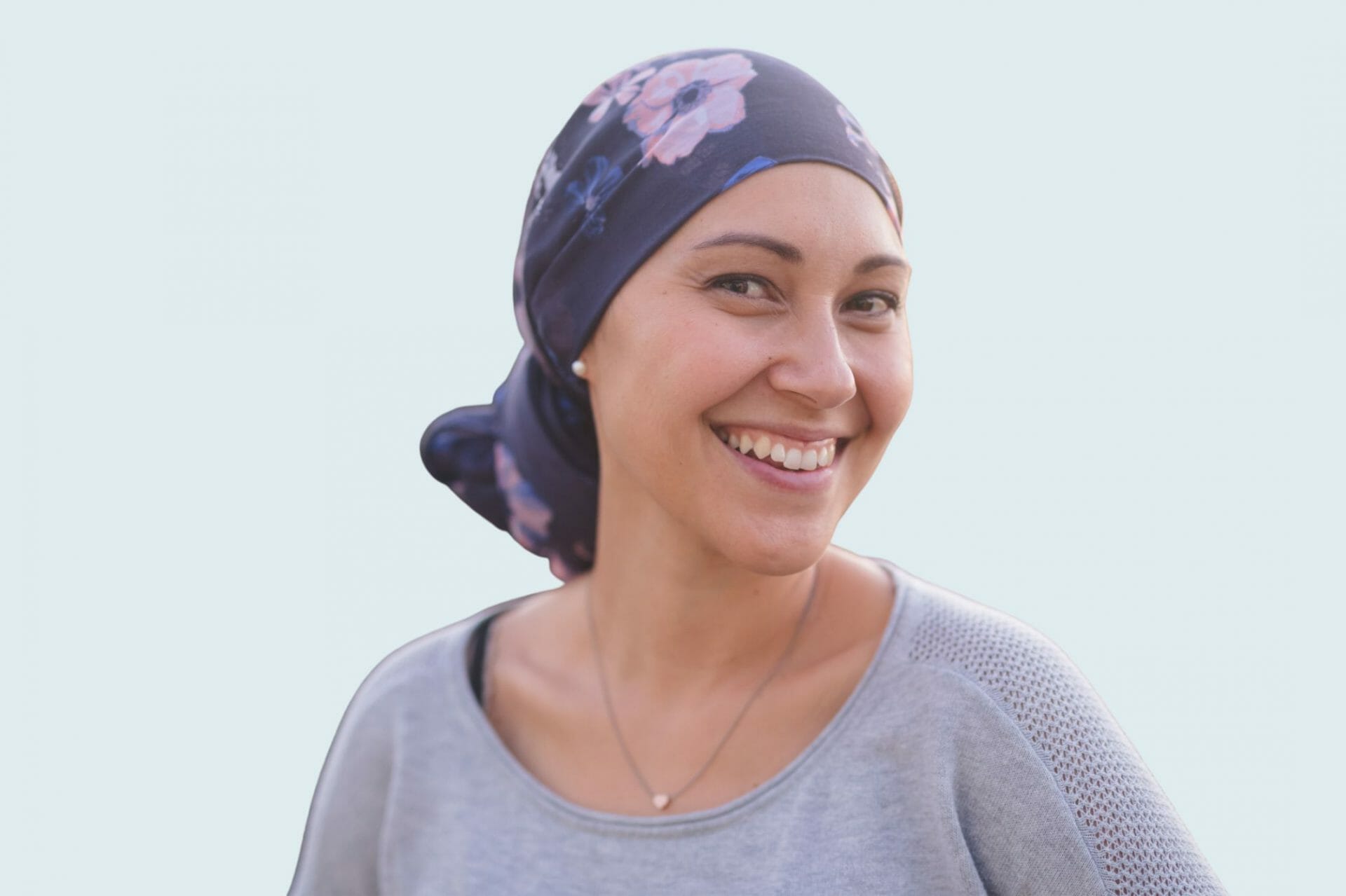 A smiling woman wearing a floral headscarf and a gray sweater. She appears to be a cancer patient, radiating warmth and positivity. The background is a soft, light blue, creating a calm and uplifting atmosphere.