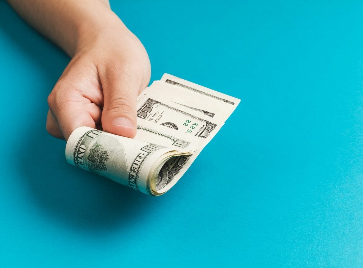 Hand holding a roll of dollar bills against a blue background, symbolizing financial assistance for cancer patients in Illinois.