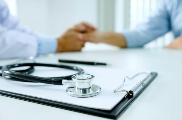 A stethoscope rests on a clipboard in the foreground, while in the background, a doctor holds a patient's hands, offering comfort and support. The setting is a bright, professional medical office.